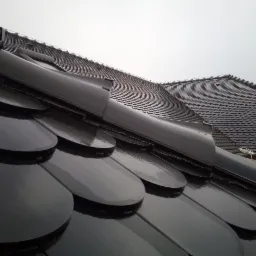 Błyszczące, czarne dachówki ceramiczne w kształcie rybiej łuski, widok z dołu pod kątem, z elementami wentylacyjnymi i fragmentem drugiego dachu w tle, pochmurne niebo.