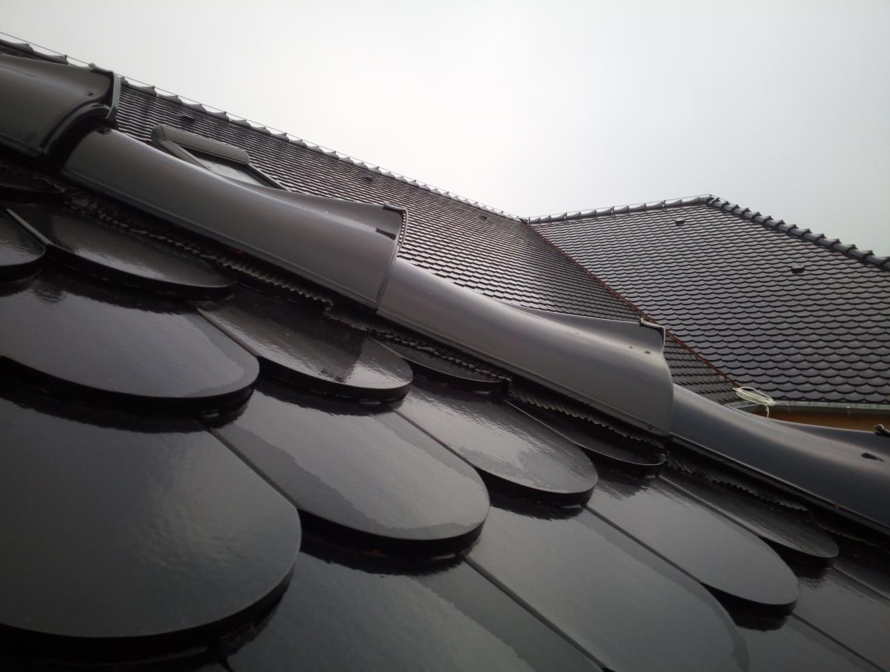 Błyszczące, czarne dachówki ceramiczne w kształcie rybiej łuski, widok z dołu pod kątem, z elementami wentylacyjnymi i fragmentem drugiego dachu w tle, pochmurne niebo.