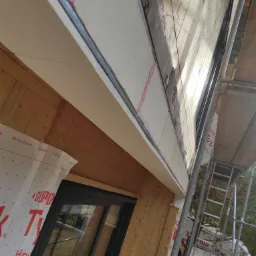 Fragment elewacji domu w trakcie montażu: widoczne drewniane panele, warstwa izolacyjna z różową pianką, membrana wiatroizolacyjna DuPont Tyvek oraz rusztowanie.