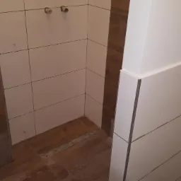 Fragment łazienki w trakcie prac wykończeniowych: ułożone brązowe płytki podłogowe imitujące drewno, białe płytki ścienne z szarą fugą, widoczne przyłącza hydrauliczne i stelaż podtynkowy WC.