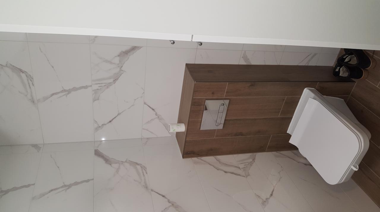 Nowoczesna toaleta podwieszana z zabudową w kolorze drewna, białe płytki imitujące marmur na ścianach i podłodze, widoczne buty w rogu.