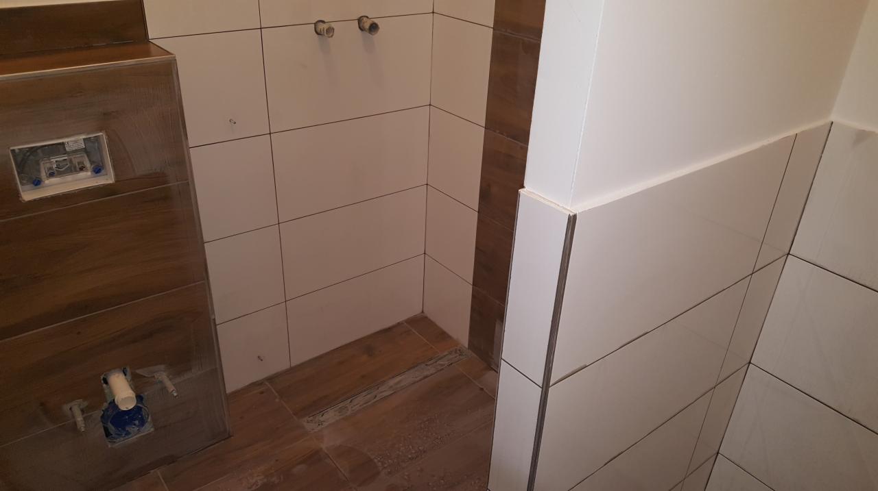 Fragment łazienki w trakcie prac wykończeniowych: ułożone brązowe płytki podłogowe imitujące drewno, białe płytki ścienne z szarą fugą, widoczne przyłącza hydrauliczne i stelaż podtynkowy WC.