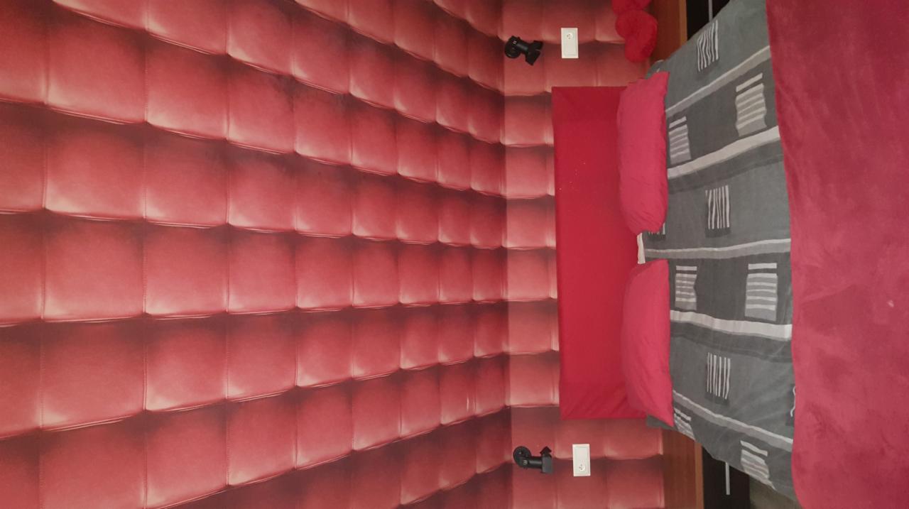 Czerwona ściana z tapetą imitującą pikowaną skórę, czarne reflektory ścienne i fragment łóżka z szarą pościelą w pokoju.