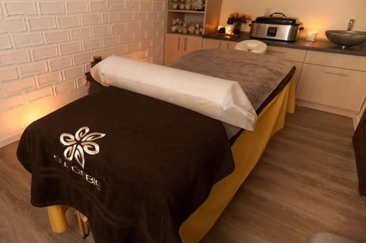 Przytulne wnętrze gabinetu kosmetycznego z profesjonalnym łóżkiem do masażu, przykrytym brązowym pokrowcem z logo i białym wałkiem pod głowę, w tle biała ceglana ściana i szafki z dekoracjami.