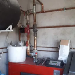 Czerwony piec CO z czarnymi drzwiczkami, widoczne miedziane rury, białe wiadro i plamy wilgoci na suficie w pomieszczeniu.