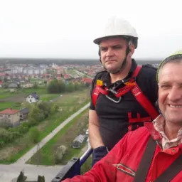 Dwóch mężczyzn w kaskach i uprzężach na wysokości z widokiem na miasto i pola, jeden uśmiechnięty, drugi z poważną miną.