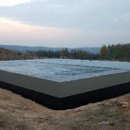 Wylana płyta fundamentowa z izolacją przeciwwilgociową, widoczna osoba w oddali i fragment budowanego domu w tle.