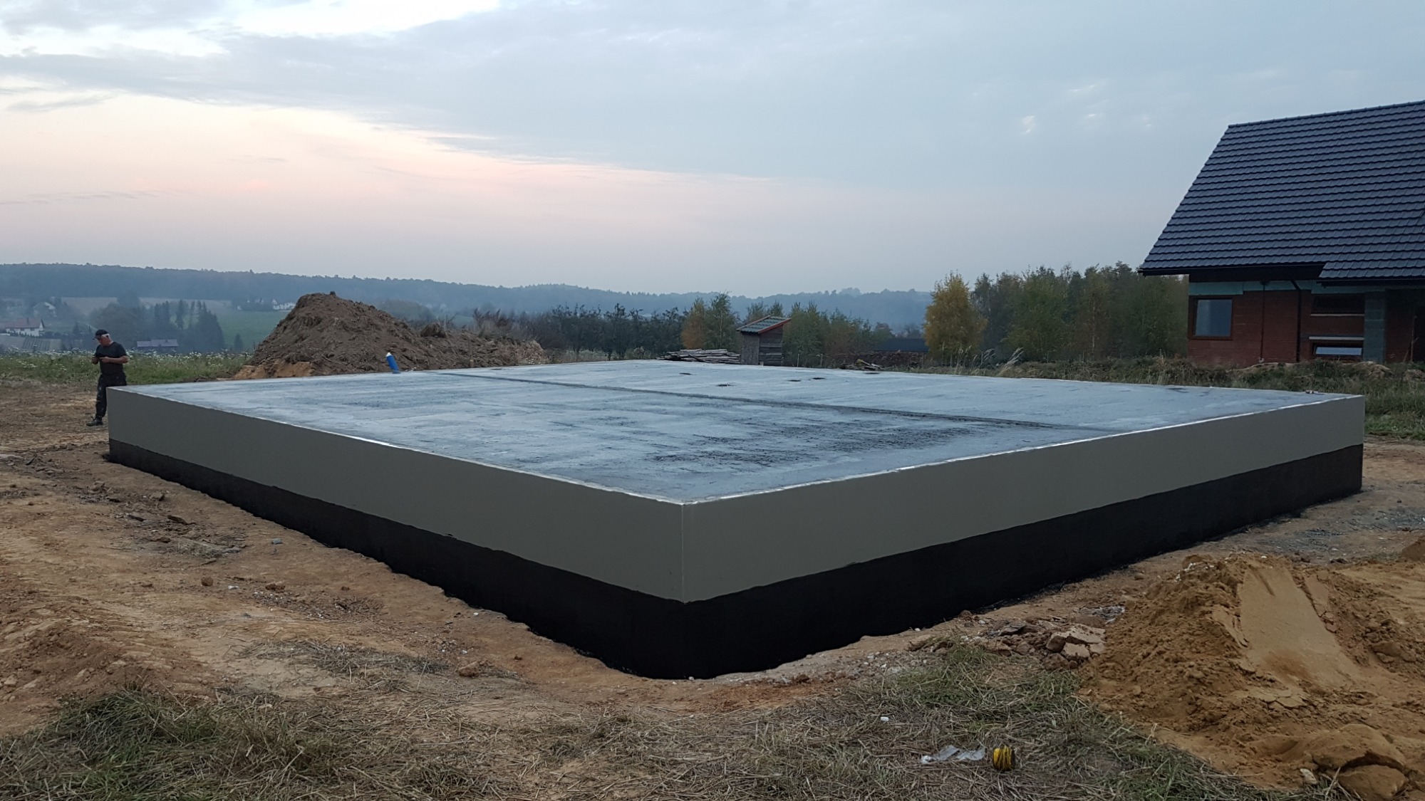 Wylana płyta fundamentowa z izolacją przeciwwilgociową, widoczna osoba w oddali i fragment budowanego domu w tle.