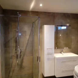 Nowoczesna łazienka z kabiną prysznicową ze szklanymi drzwiami, białą szafką łazienkową z umywalką i lustrem, wykończona brązowymi płytkami.