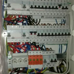 Otwarta skrzynka rozdzielcza z bezpiecznikami, okablowaniem w kolorach czerwonym, niebieskim, zielonym i żółtym, listwa zaciskowa u dołu, widoczne elementy marki ETI.