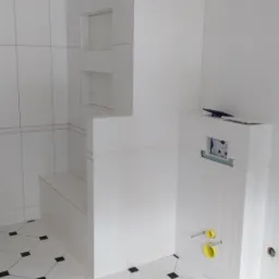 Biała łazienka w trakcie wykończenia z zabudową WC, płytkami podłogowymi w biało-czarną szachownicę, widoczne żółte rury doprowadzające wodę i elementy montażowe.