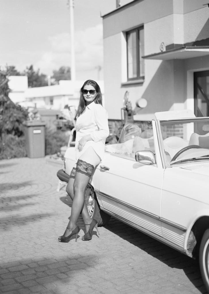 Czarno-białe zdjęcie kobiety w okularach przeciwsłonecznych, koszuli i pończochach z koronką, opierającej się o biały kabriolet na tle domu.