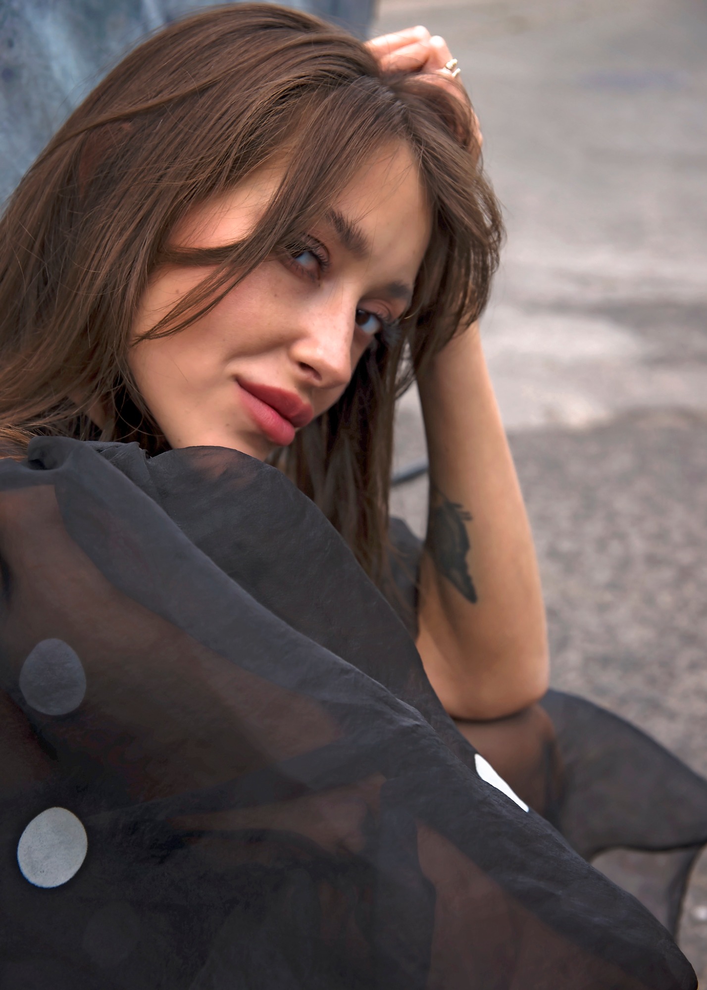 Portret kobiety z brązowymi włosami, w czarnej, przezroczystej bluzce w białe grochy, z tatuażem na ramieniu, opierającej głowę na dłoni z pierścionkiem.