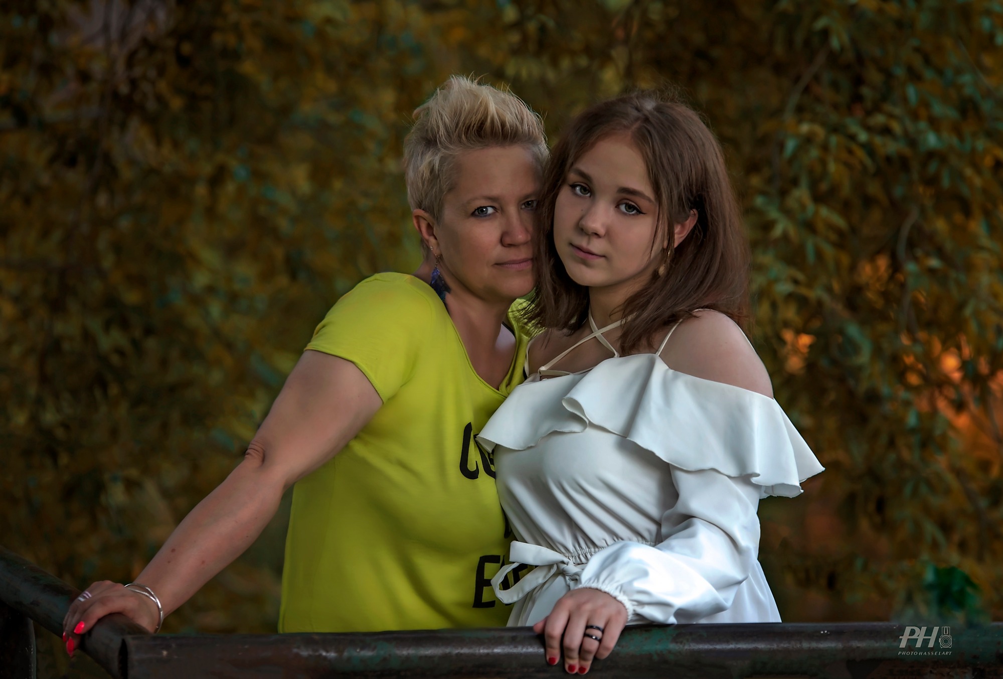 Portret matki z nastoletnią córką, obie oparte o metalową barierkę na tle jesiennych drzew, córka w białej sukience z falbanami, matka w limonkowej koszulce.