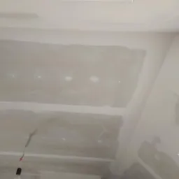 Niedokończony sufit z widocznymi łączeniami płyt gipsowo-kartonowych, z prowizorycznym oświetleniem w postaci wiszącej żarówki na kablu.