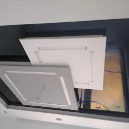 Czarny, podwieszany sufit z dwoma kwadratowymi panelami oświetleniowymi w kolorze białym i szarym, widoczne przewody elektryczne.