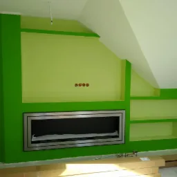 Ściana w odcieniach zieleni z wnęką na kominek obudowaną metalową ramą, widoczne gniazdka elektryczne, fragment poddasza i kartony na podłodze.