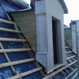 Mirosław Poszelężny - W trakcie budowy dachu: widoczne drewniane krokwie, czarna membrana dachowa z nadrukiem, surowa lukarna z szarego betonu oraz komin w tle.