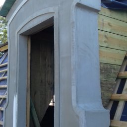 Mirosław Poszelężny - Wznoszenie lukarny dachowej z surowym wykończeniem tynkarskim, widoczne drewniane elementy konstrukcji dachu i izolacja, etap prac wykończeniowych.