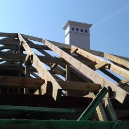 Mirosław Poszelężny - Drewniana konstrukcja dachu w trakcie budowy, widoczne krokwie i więźba dachowa, z kominem w tle na tle błękitnego nieba.