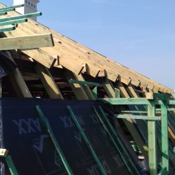 Mirosław Poszelężny - Konstrukcja drewniana dachu w trakcie budowy, widoczna membrana dachowa z nadrukiem, błękitne niebo w tle.