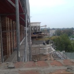 Mirosław Poszelężny - Renowacja zabytkowej wieży z cegły, widok z rusztowania na ekipę pracującą na górze, w tle zabudowa miejska i zieleń.