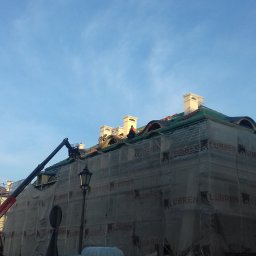 Mirosław Poszelężny - Budynek w trakcie prac dekarskich, zabezpieczony siatką rusztowaniową z widocznym dźwigiem i pracownikami na dachu, błękitne niebo w tle.
