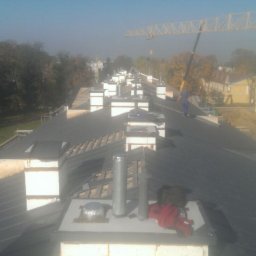 Mirosław Poszelężny - Długi, blaszany dach z licznymi kominami w trakcie montażu, widoczny dźwig budowlany w tle, narzędzia i ubrania robocze na kominie.