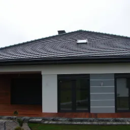 Nowoczesny dom z ciemnoszarą dachówką ceramiczną, widoczne okno dachowe i komin, drewniany taras oraz okna z roletami zewnętrznymi.