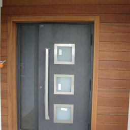 Mirosław Poszelężny - Nowoczesne, grafitowe drzwi wejściowe z trzema kwadratowymi, matowymi przeszkleniami i długim, srebrnym uchwytem, otoczone drewnianą ramą i wykończone drewnianą elewacją.
