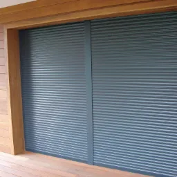 Zewnętrzne rolety antywłamaniowe w kolorze grafitowym, zamontowane na elewacji obłożonej drewnianymi panelami, widoczne z poziomu tarasu.