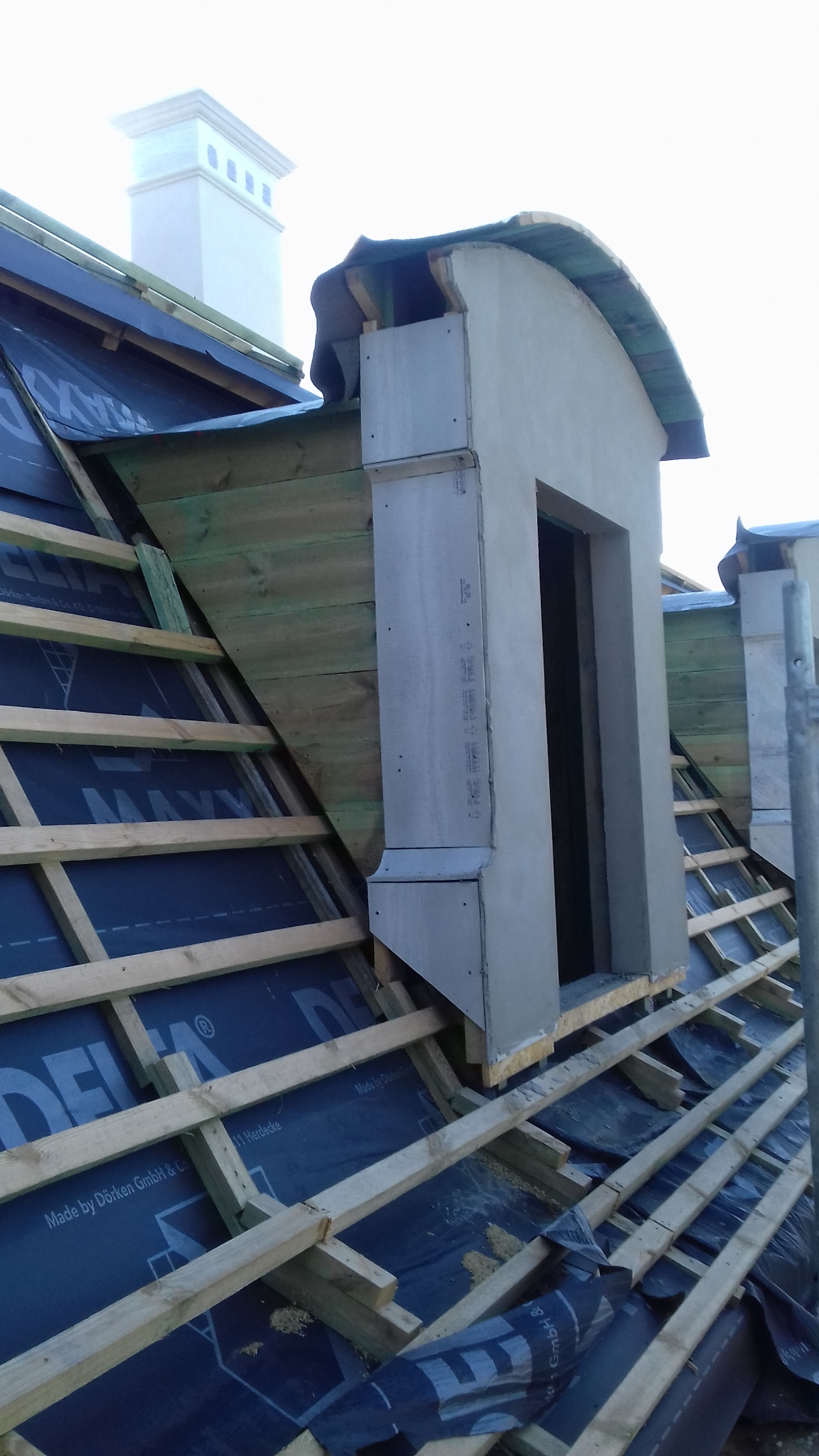 W trakcie budowy dachu: widoczne drewniane krokwie, czarna membrana dachowa z nadrukiem, surowa lukarna z szarego betonu oraz komin w tle.