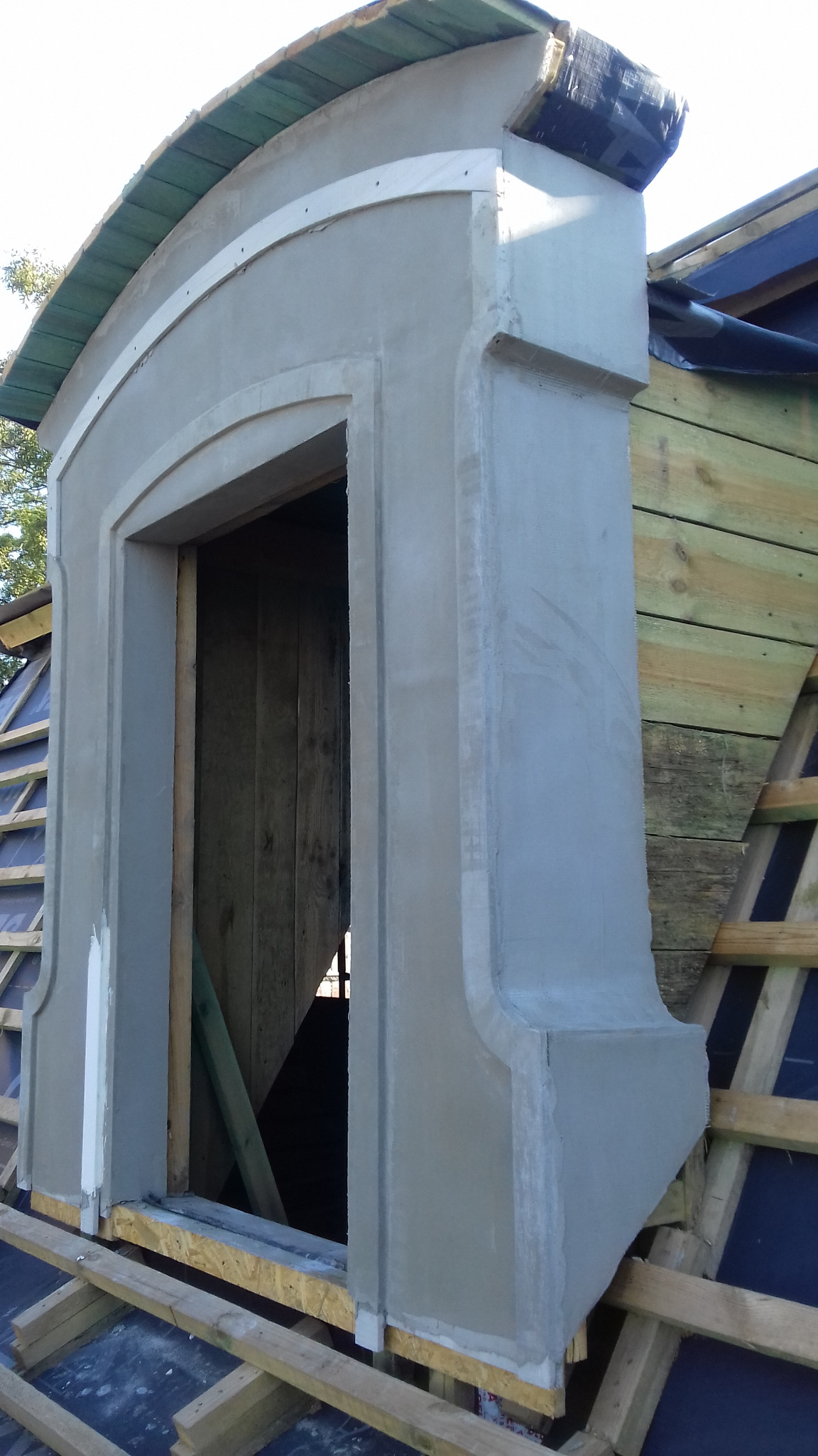 Wznoszenie lukarny dachowej z surowym wykończeniem tynkarskim, widoczne drewniane elementy konstrukcji dachu i izolacja, etap prac wykończeniowych.