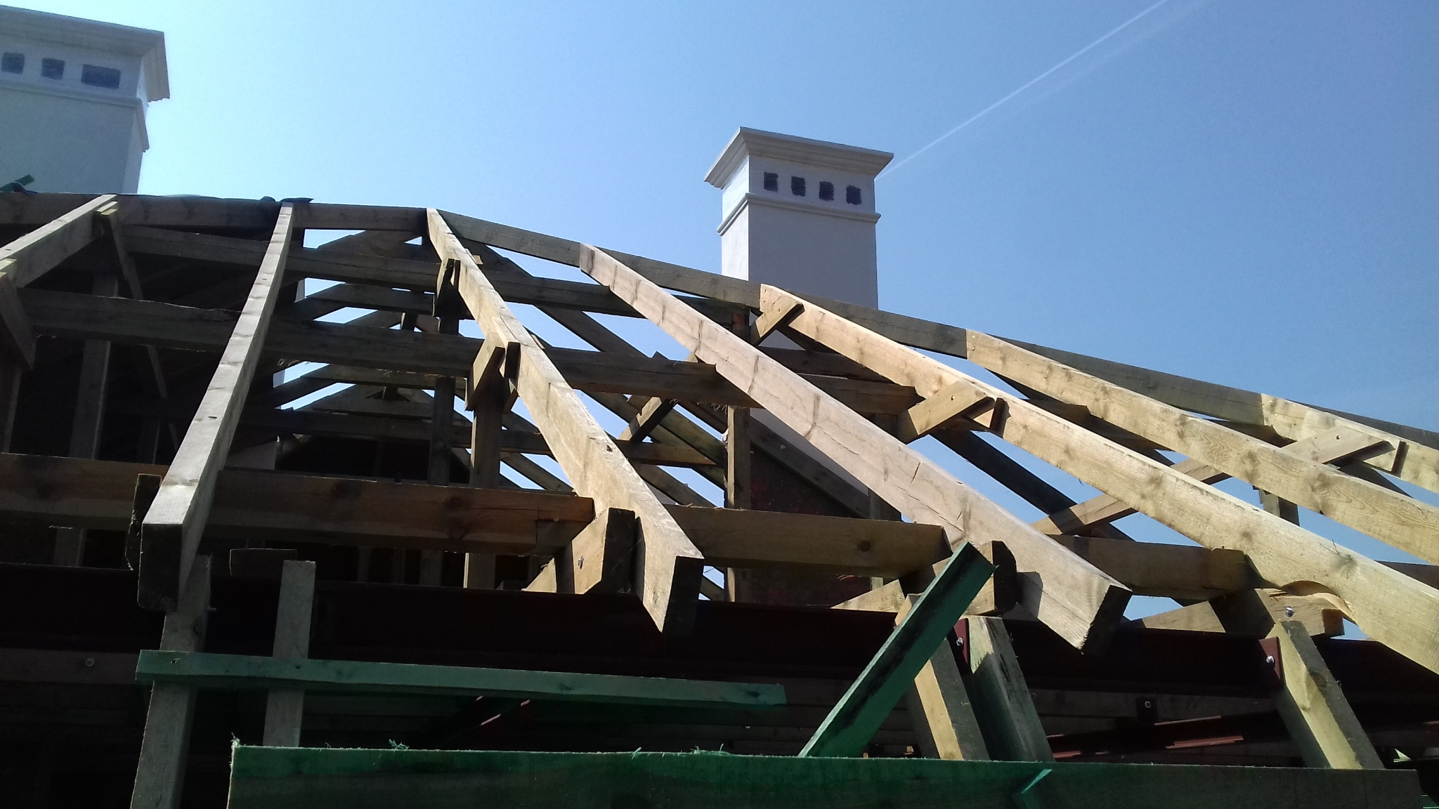 Drewniana konstrukcja dachu w trakcie budowy, widoczne krokwie i więźba dachowa, z kominem w tle na tle błękitnego nieba.