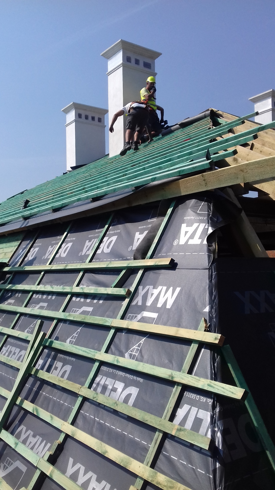 Ekipa dekarzy w trakcie montażu kontrłat na folii paroizolacyjnej dachu, widoczne kominy w tle.