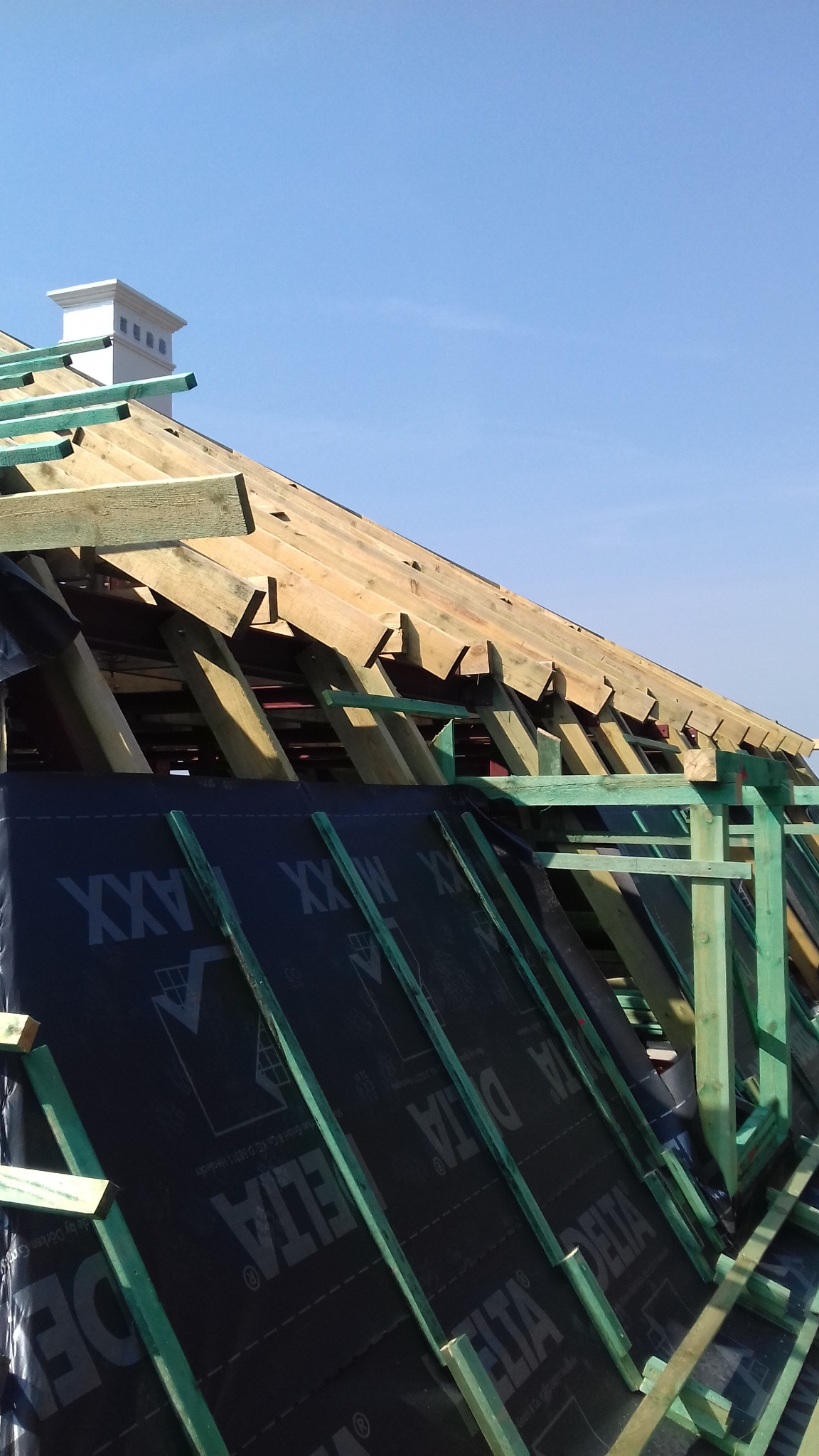 Konstrukcja drewniana dachu w trakcie budowy, widoczna membrana dachowa z nadrukiem, błękitne niebo w tle.