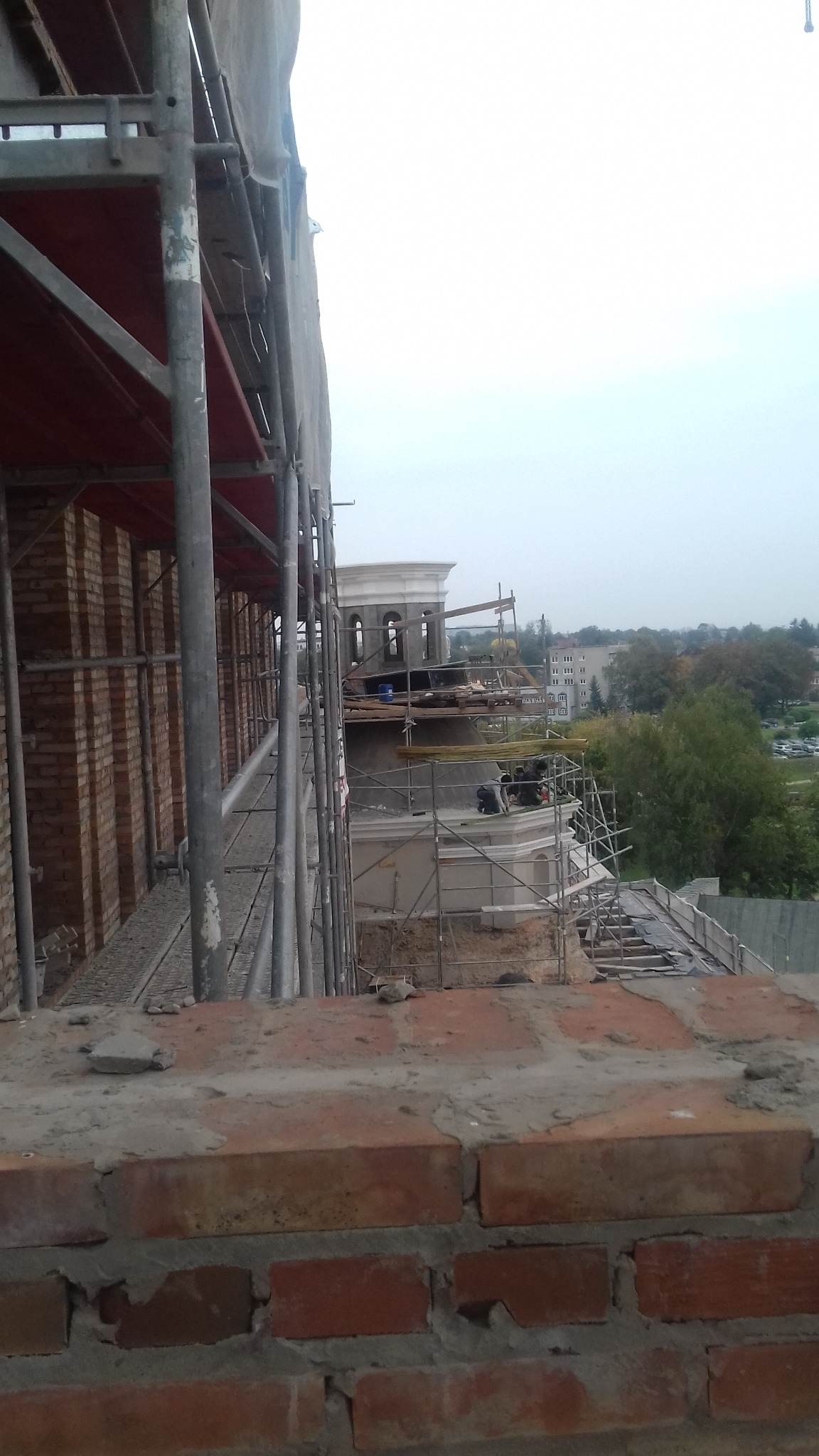 Renowacja zabytkowej wieży z cegły, widok z rusztowania na ekipę pracującą na górze, w tle zabudowa miejska i zieleń.