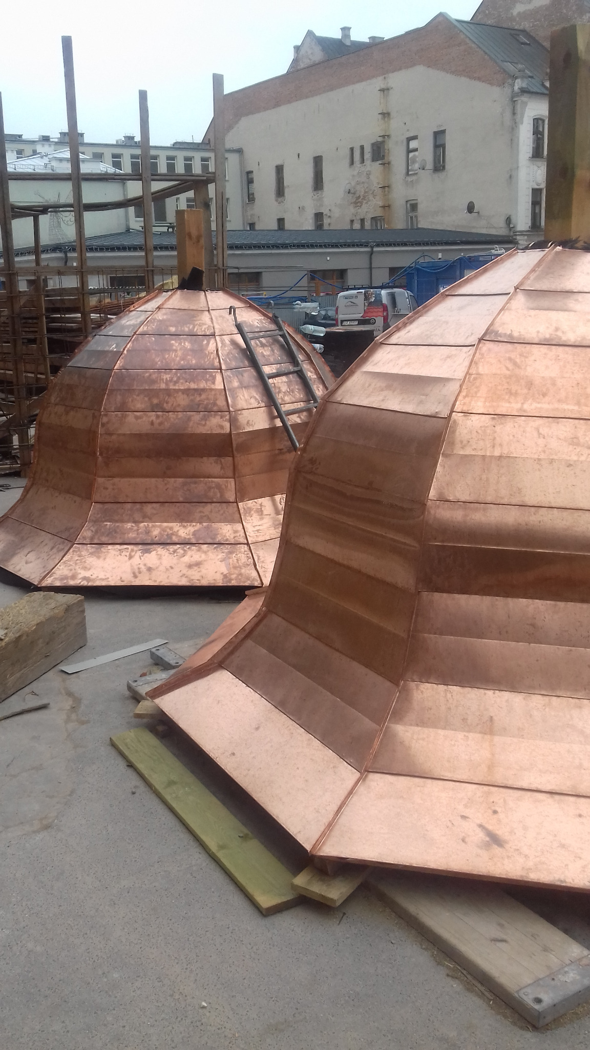 Dwa miedziane, kopulaste elementy konstrukcyjne, prawdopodobnie wieże, w trakcie budowy lub renowacji, z widoczną drabiną opartą o jeden z nich, na tle miejskiego krajobrazu z budynkami...