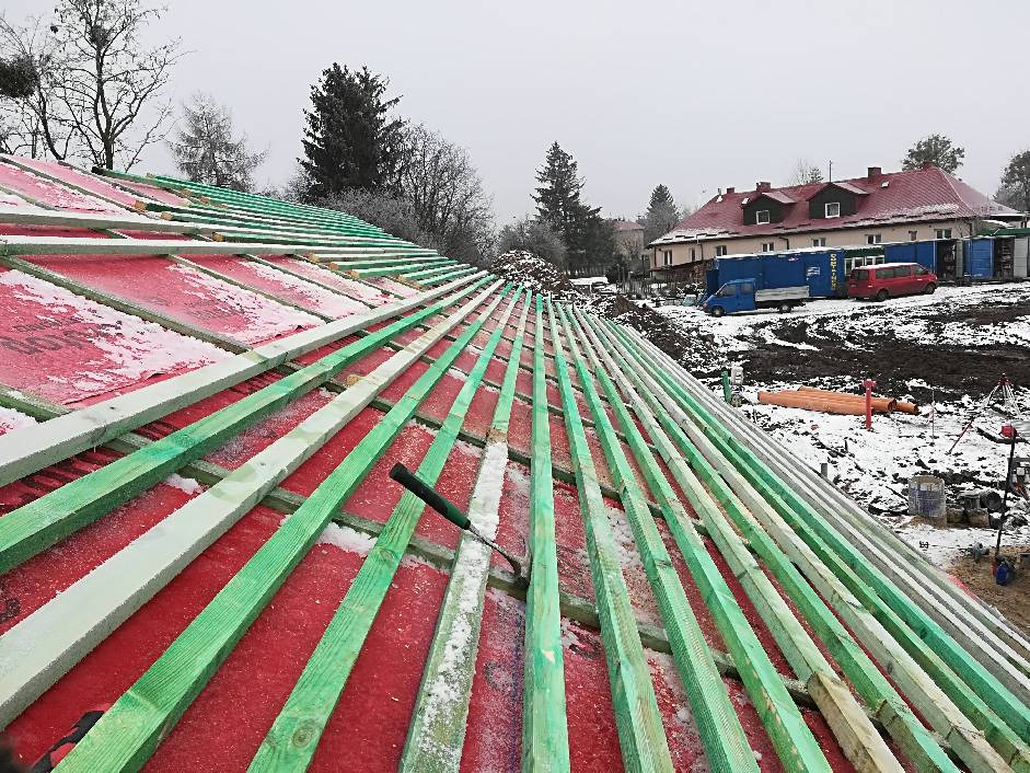 Konstrukcja dachu z zielonymi łatami na czerwonej membranie, pokryta warstwą świeżego śniegu, w tle budynek i teren budowy.