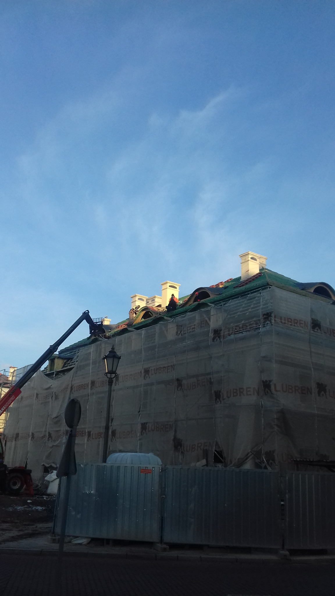 Budynek w trakcie prac dekarskich, zabezpieczony siatką rusztowaniową z widocznym dźwigiem i pracownikami na dachu, błękitne niebo w tle.