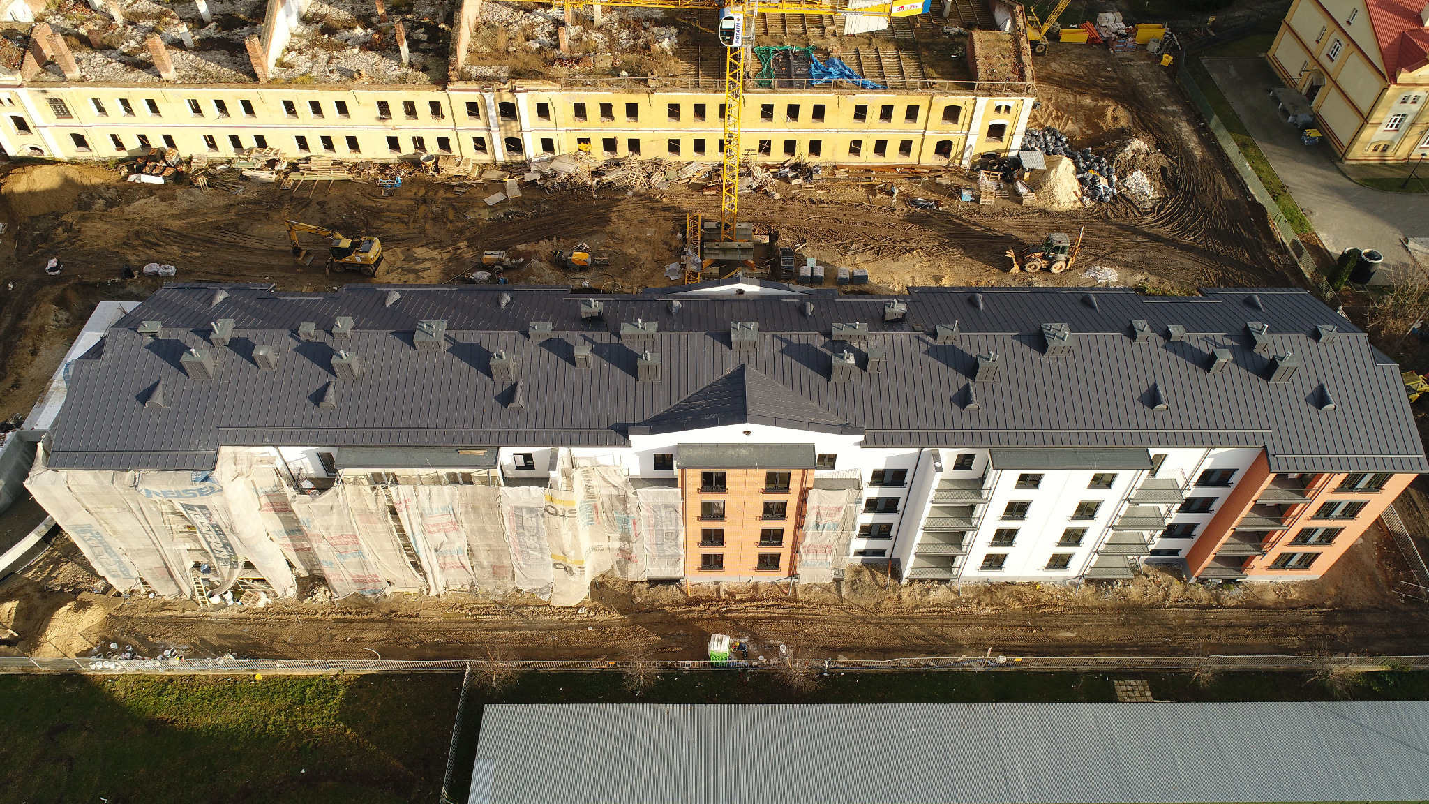 Widok z góry na budynek wielorodzinny w trakcie budowy z nowym, ciemnoszarym dachem blaszanym, częściowo osłonięty rusztowaniami i siatkami ochronnymi, w tle plac budowy z żółtym dźwigiem i stary...