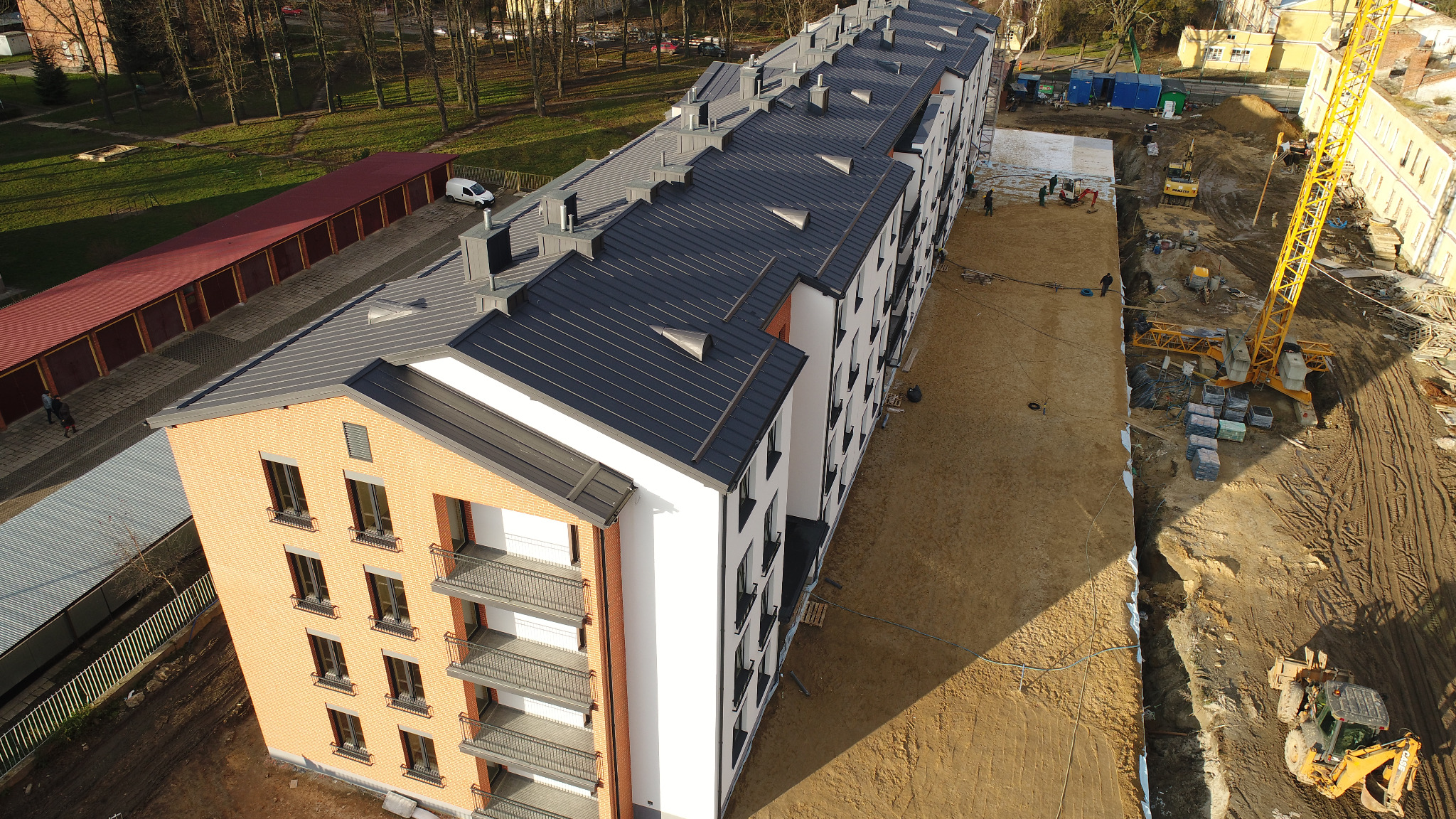 Widok z góry na nowo wybudowany, wielopiętrowy budynek mieszkalny z ciemnoszarym dachem i balkonami, otoczony placem budowy z ciężkim sprzętem i materiałami budowlanymi.