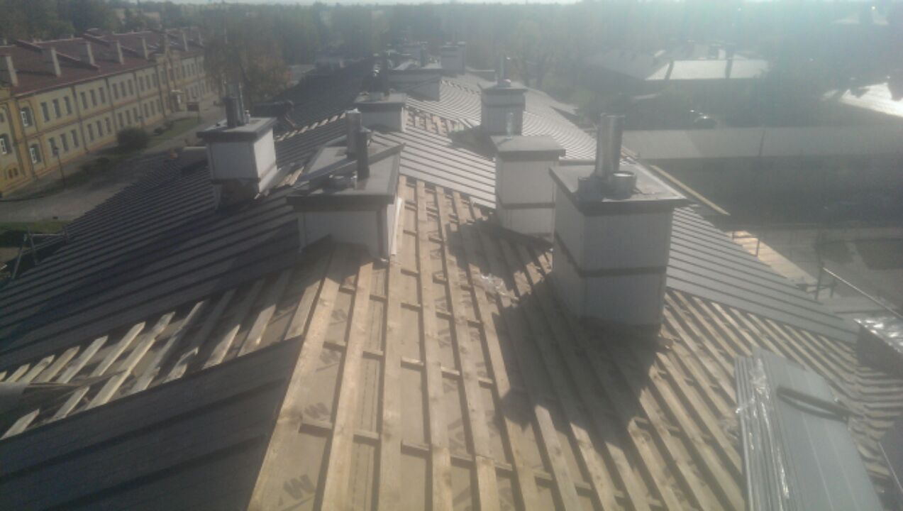 Dach z blachy trapezowej w trakcie montażu, widoczne drewniane łaty, kominy i budynek w tle, ujęcie z góry.