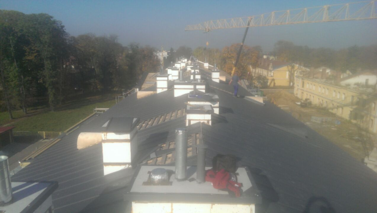 Długi, blaszany dach z licznymi kominami w trakcie montażu, widoczny dźwig budowlany w tle, narzędzia i ubrania robocze na kominie.