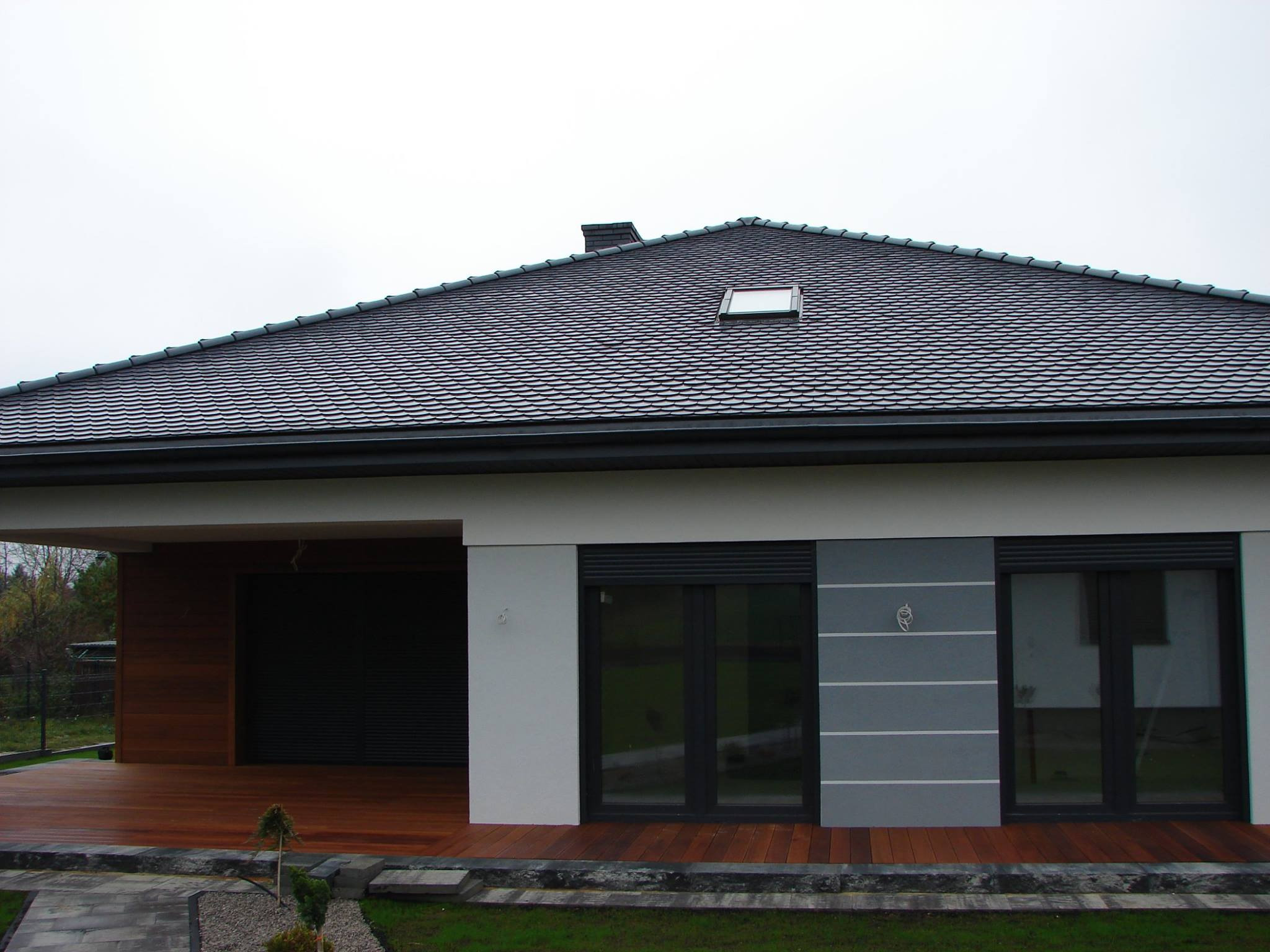 Nowoczesny dom z ciemnoszarą dachówką ceramiczną, widoczne okno dachowe i komin, drewniany taras oraz okna z roletami zewnętrznymi.