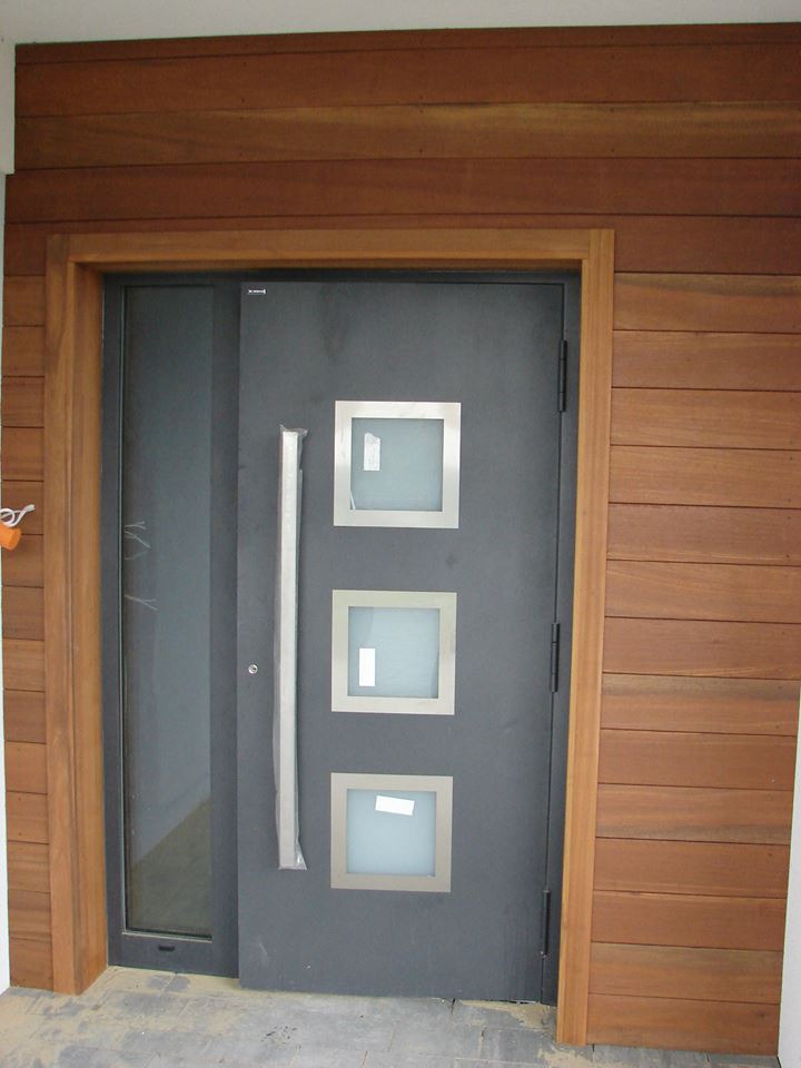 Nowoczesne, grafitowe drzwi wejściowe z trzema kwadratowymi, matowymi przeszkleniami i długim, srebrnym uchwytem, otoczone drewnianą ramą i wykończone drewnianą elewacją.