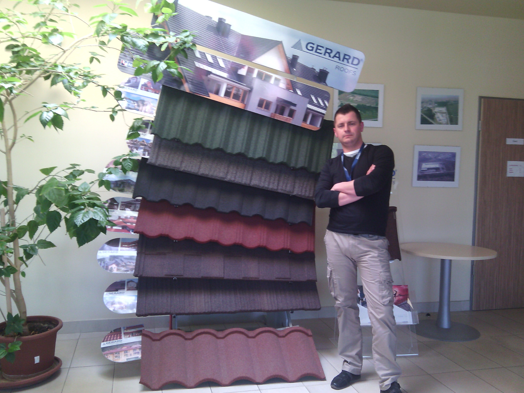 Mężczyzna prezentuje ekspozycję z różnymi kolorami i wzorami dachówek marki Gerard, obok stoi doniczka z rośliną, a na ścianie wiszą zdjęcia.