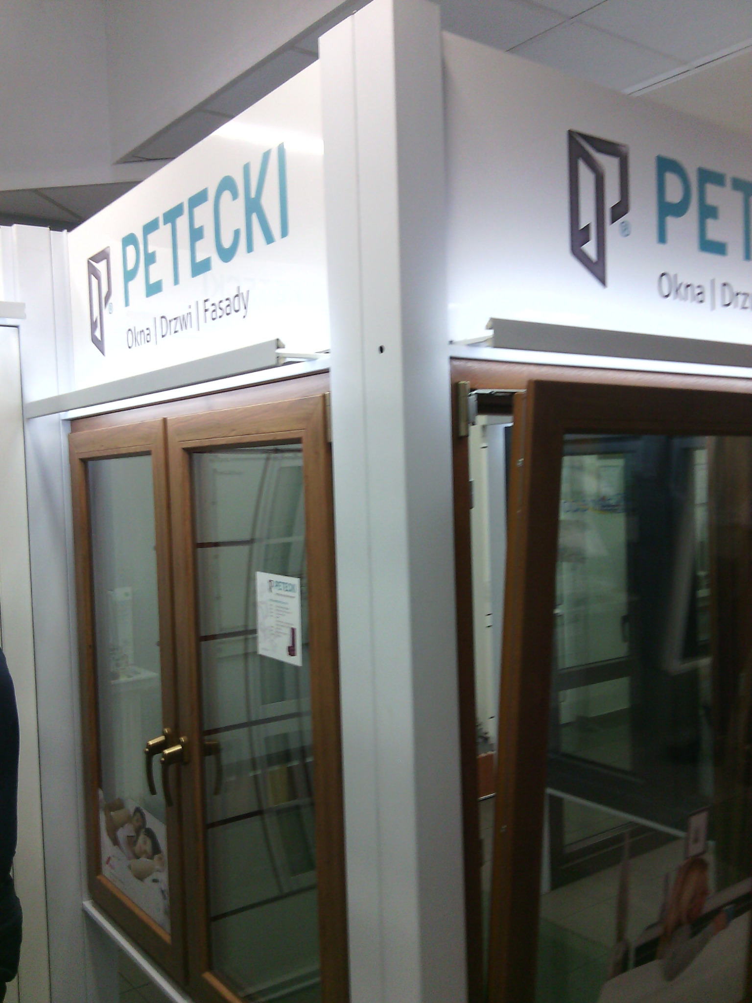Ekspozycja okien drewnianych z szybami zespolonymi w salonie firmy Petecki, widoczne logo firmy i hasło 'Okna | Drzwi | Fasady'.