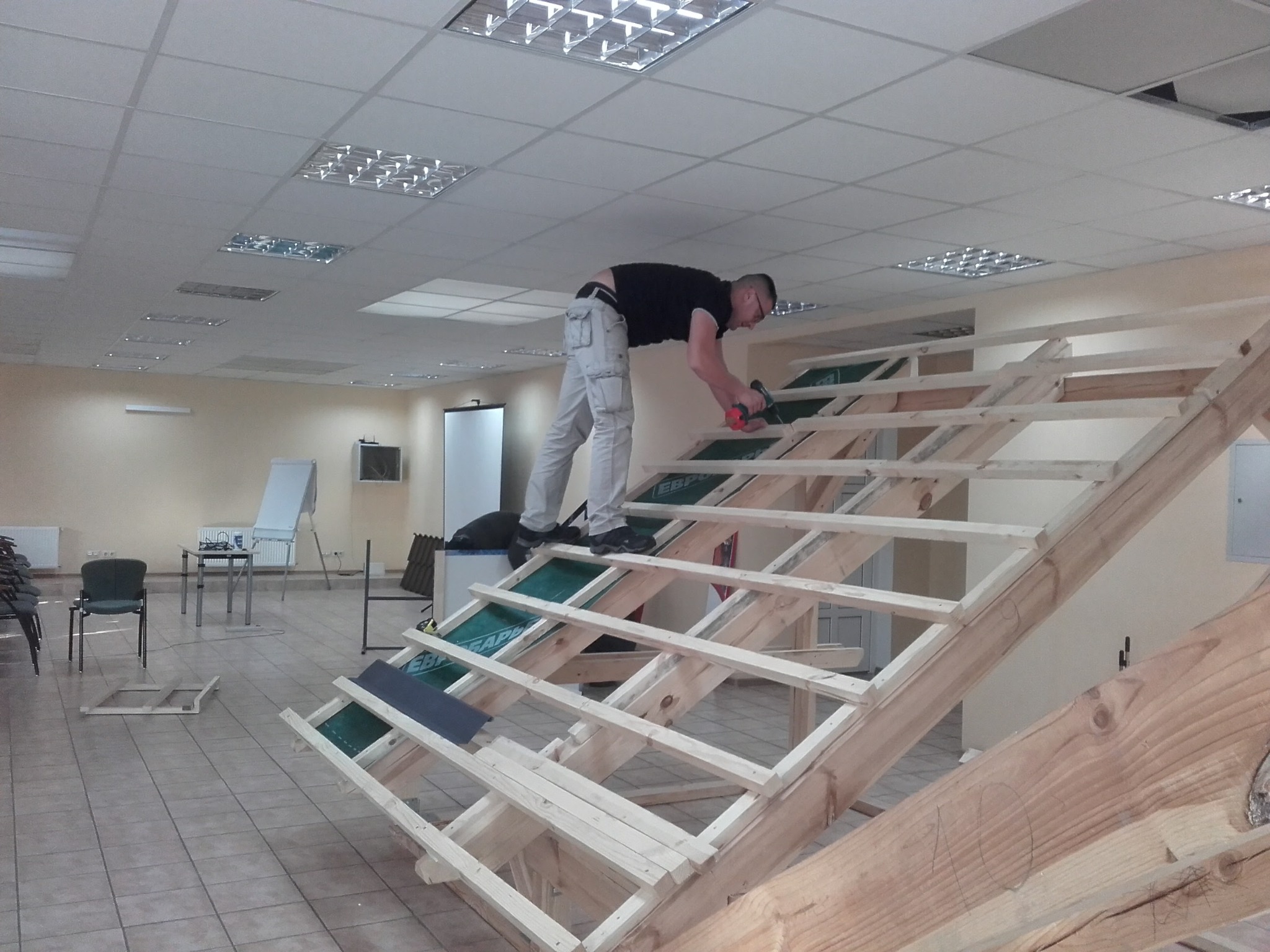 Pracownik w czarnej koszulce i jasnych spodniach montuje drewnianą konstrukcję dachu wewnątrz pomieszczenia, używając wkrętarki. Widoczne elementy izolacji i fragmenty pokrycia dachowego. W tle...