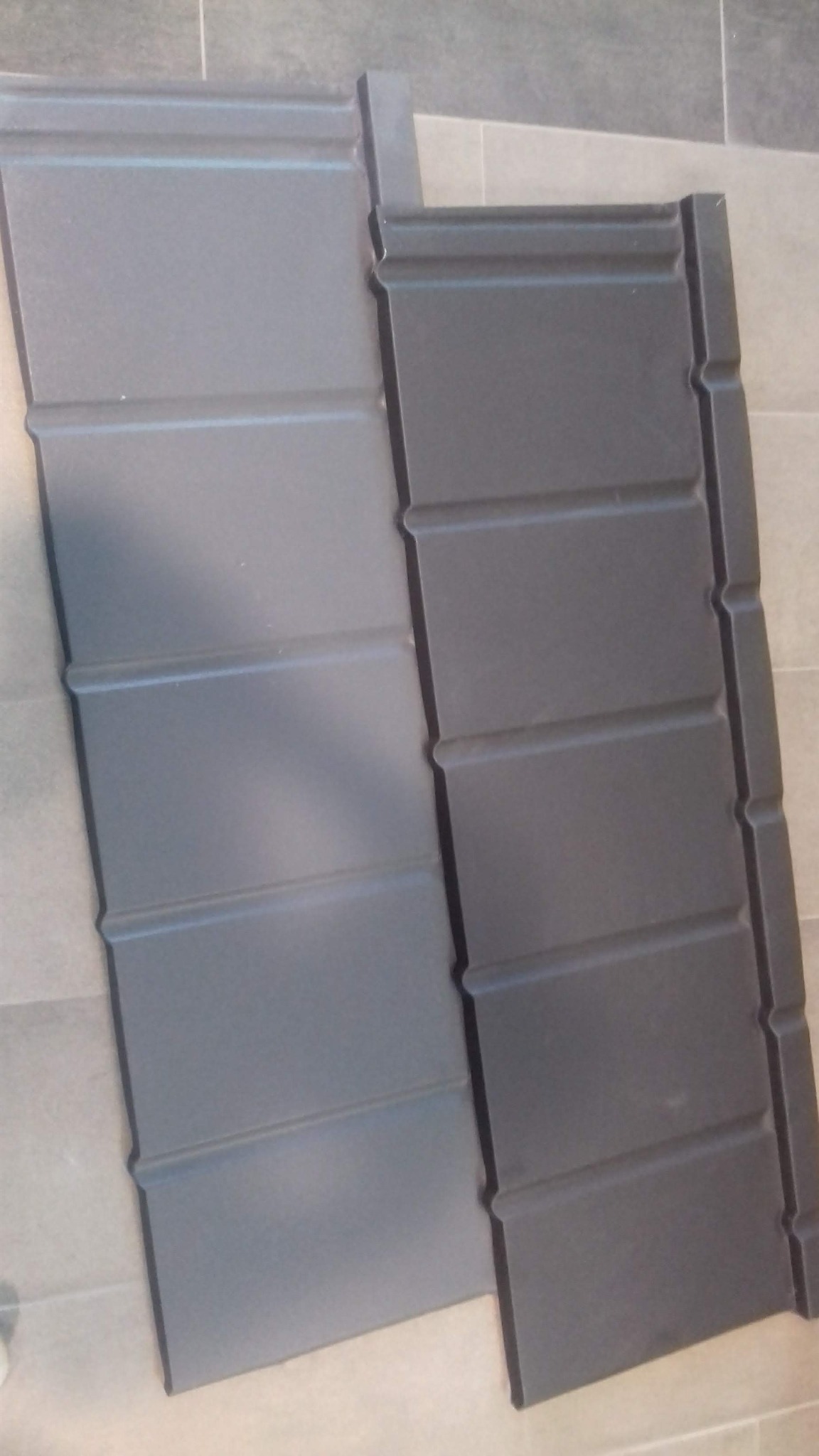 Dwa panele blaszane do pokrycia dachu, jeden w jaśniejszym odcieniu szarości, drugi ciemniejszy, oba z profilowanymi krawędziami, leżące na podłodze z płytek.